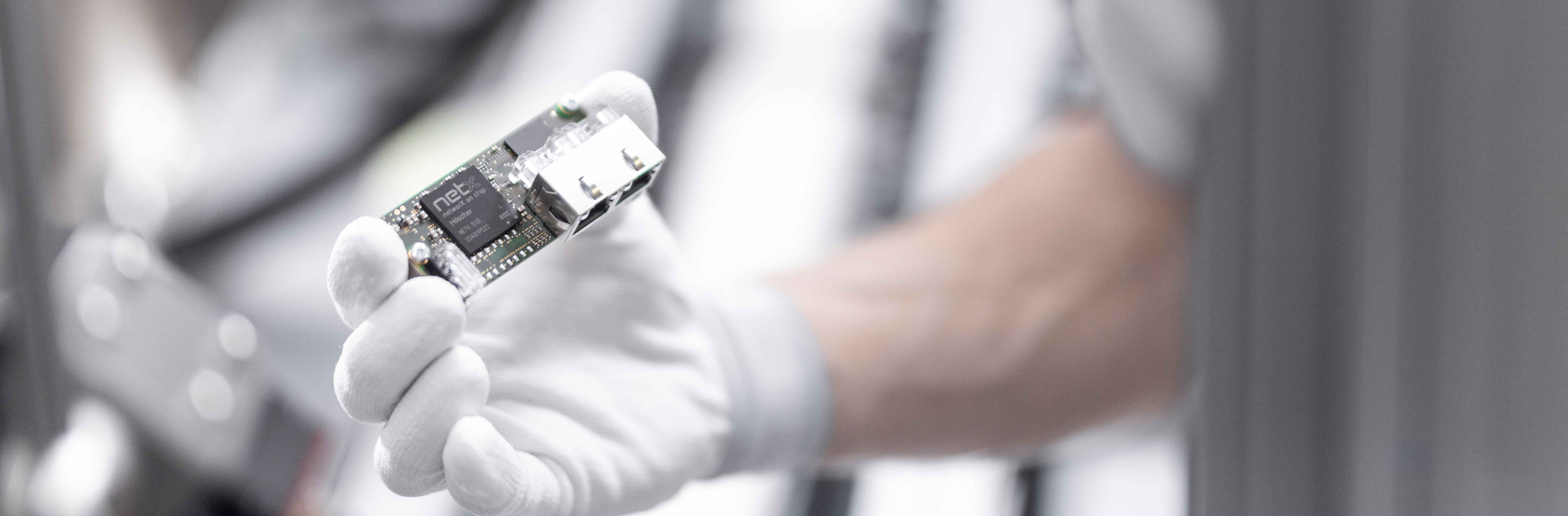 A comX module is being held by a hand in white gloves. There are two Ethernet sockets mounted on the module as well as a black netX chip. The surroundings show a semiconductor production machine and are bright and blurry.