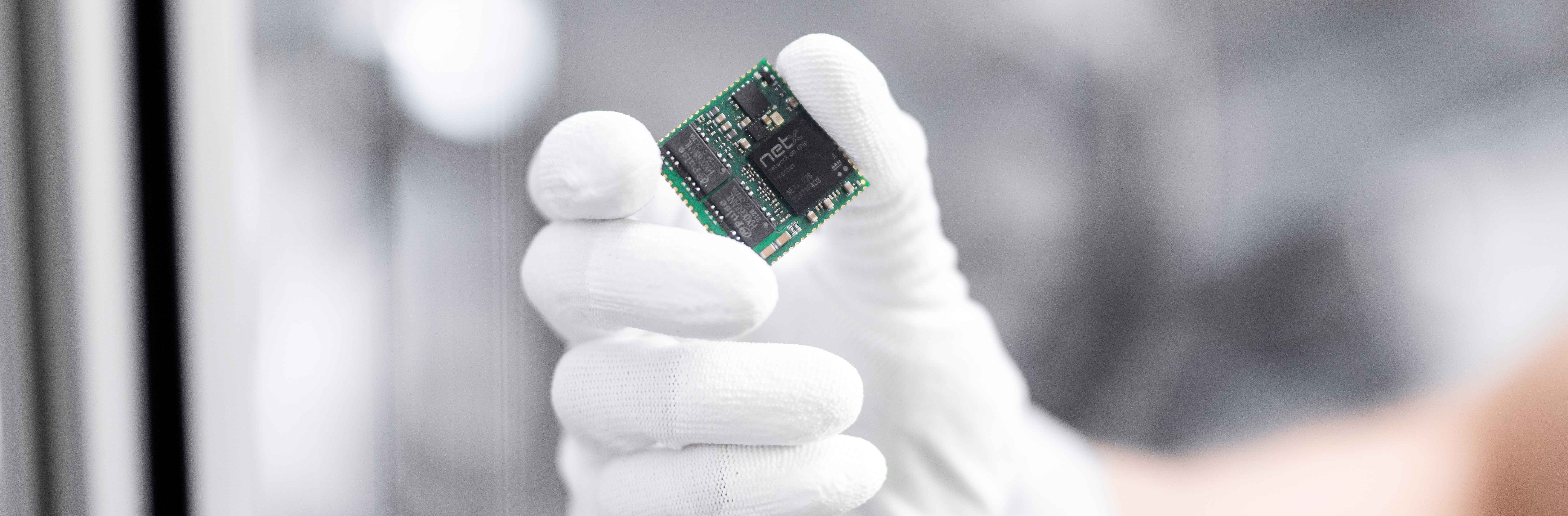 A hand in a white glove holds an embedd module from Hilscher. Mounted on the green square module is a netX 52.