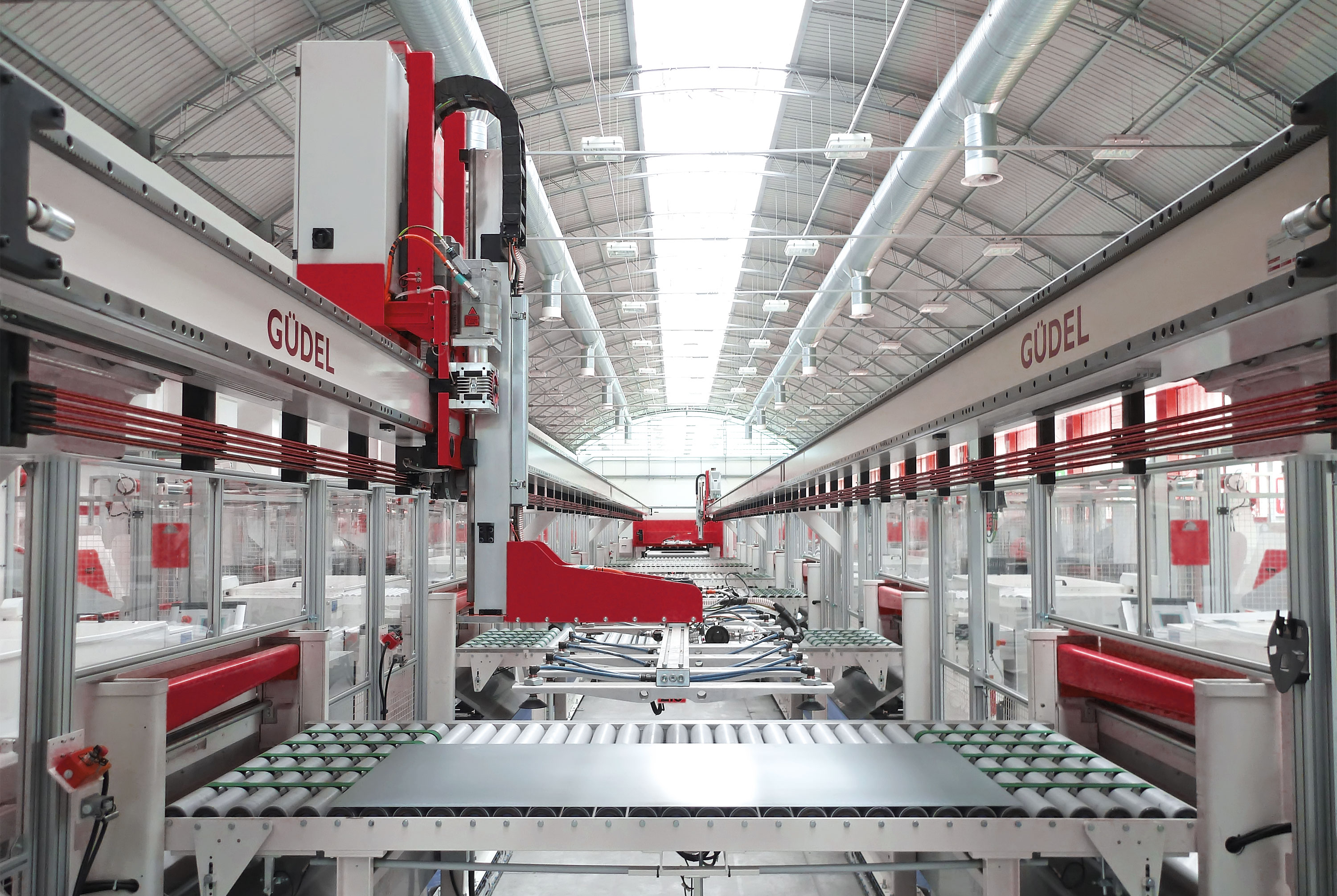 A clean and bright inside of a factory with several machines lined up left and right. On two white rails at the upper half of the picture Güdel is written in red large letters. In the foreground, a sheet of metal lays on a conveyor belt.