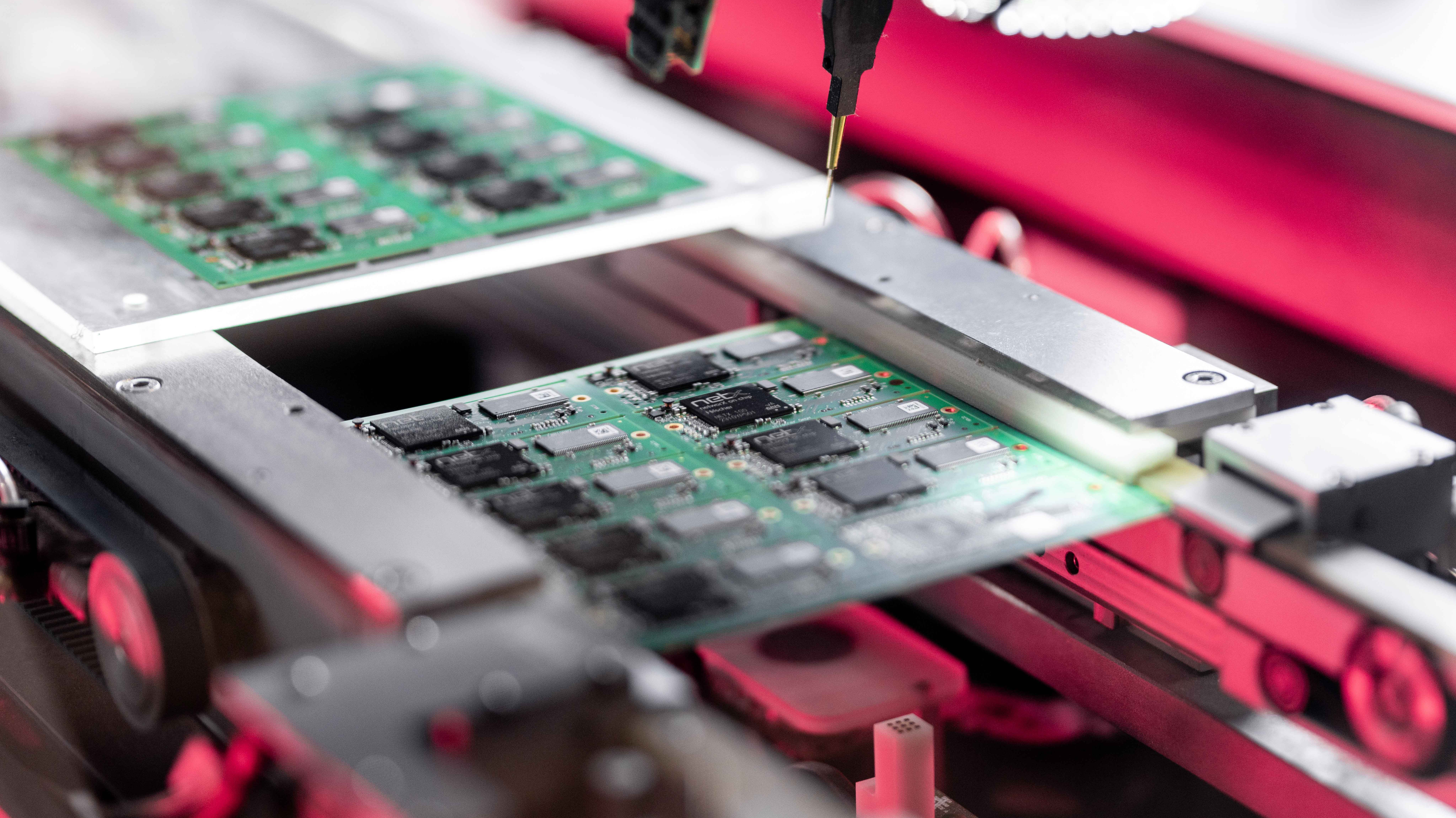 A tray of embedded modules with a netX chip onboard in a production machine. A red gleam is seen in the background. A small golden needle for testing comes from the top pointing at the tray.