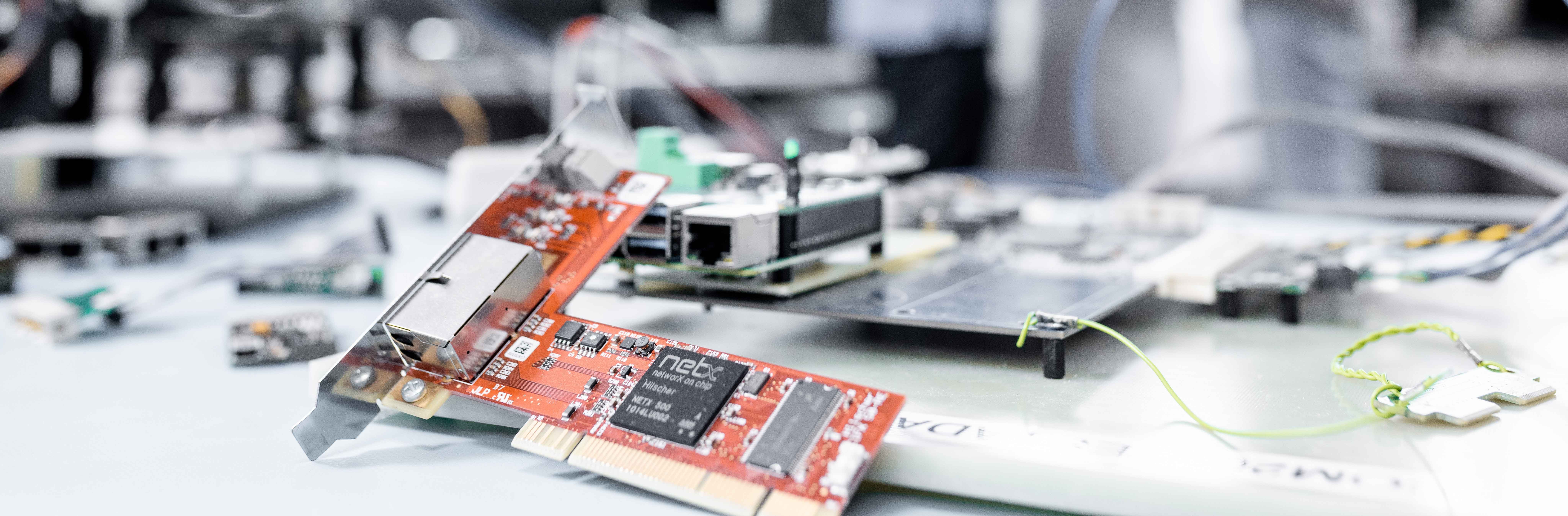 A cifX PC card with red PCB lays in the foreground surrounded by network cables and interfaces. Out of focus in the background you can see three people in a discussion 