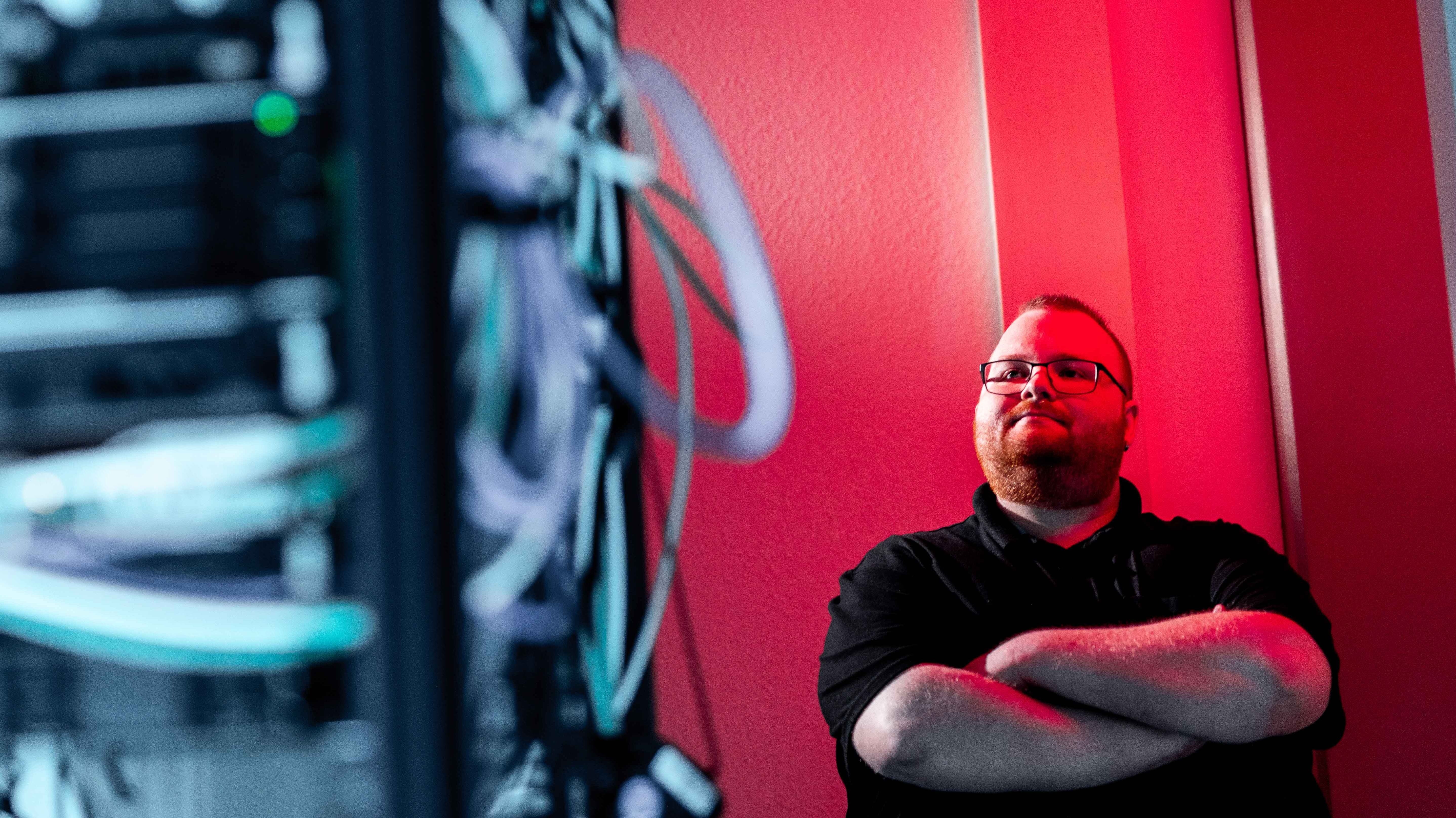 A man with glasses stands at the door of a server room, arms crossed. Red light can be seen in the background. He looks at the server racks on the left side of the picture.