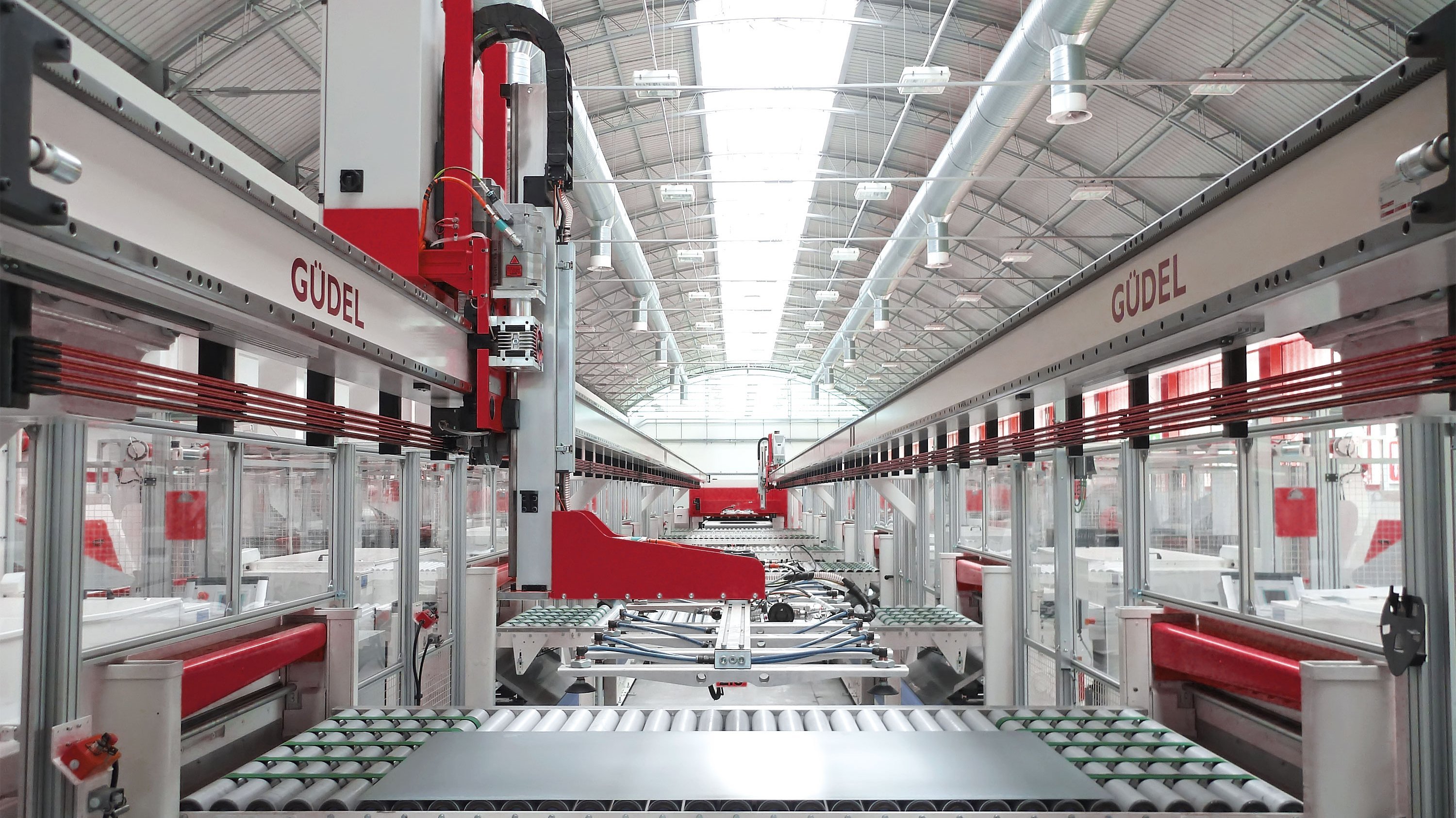 A clean and bright inside of a factory with several machines lined up left and right. On two white rails at the upper half of the picture Güdel is written in red large letters. In the foreground, a sheet of metal lays on a conveyor belt.