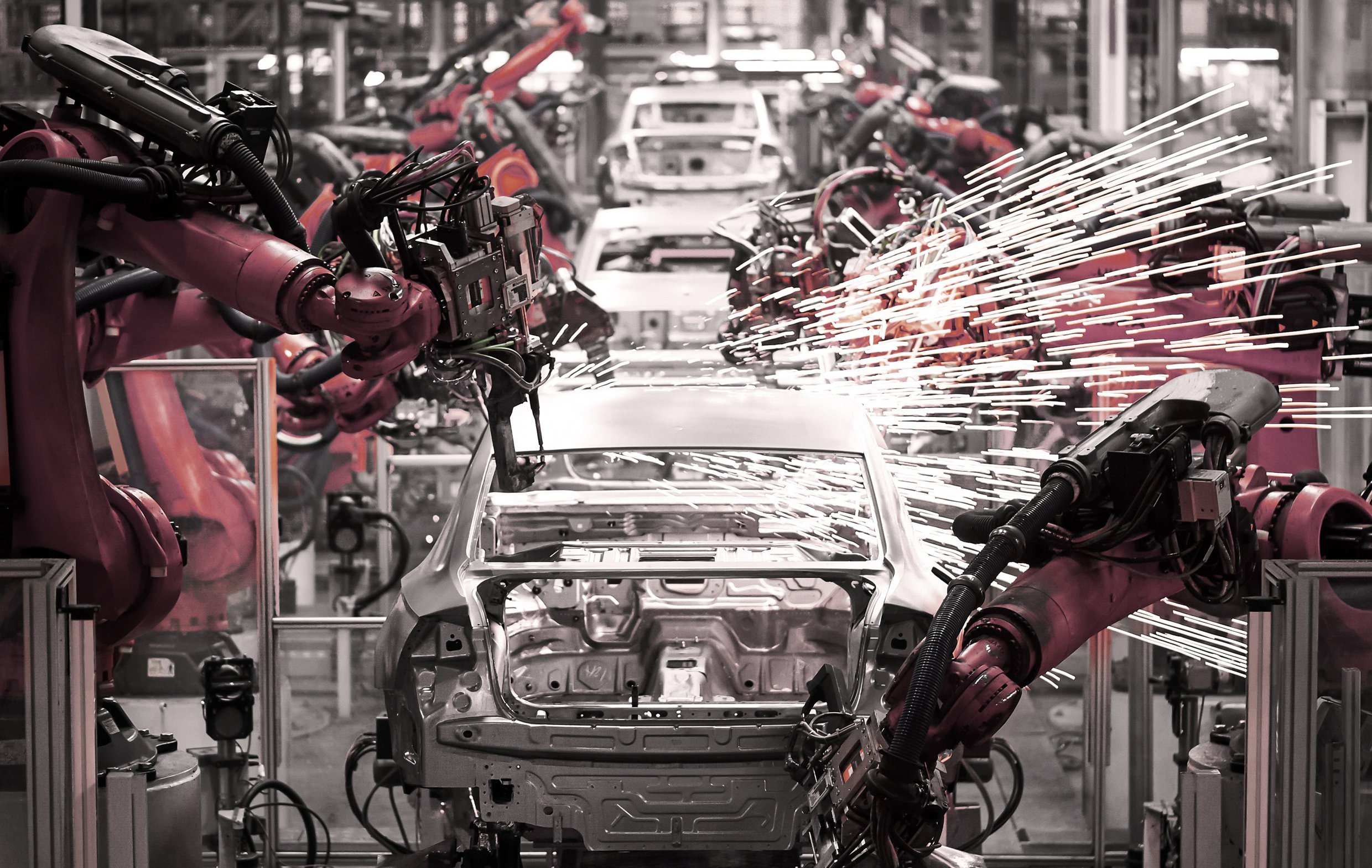 Numerous orange robots working on car chassis in an industrial production line. One robot is welding on a car creating a burst of sparks.