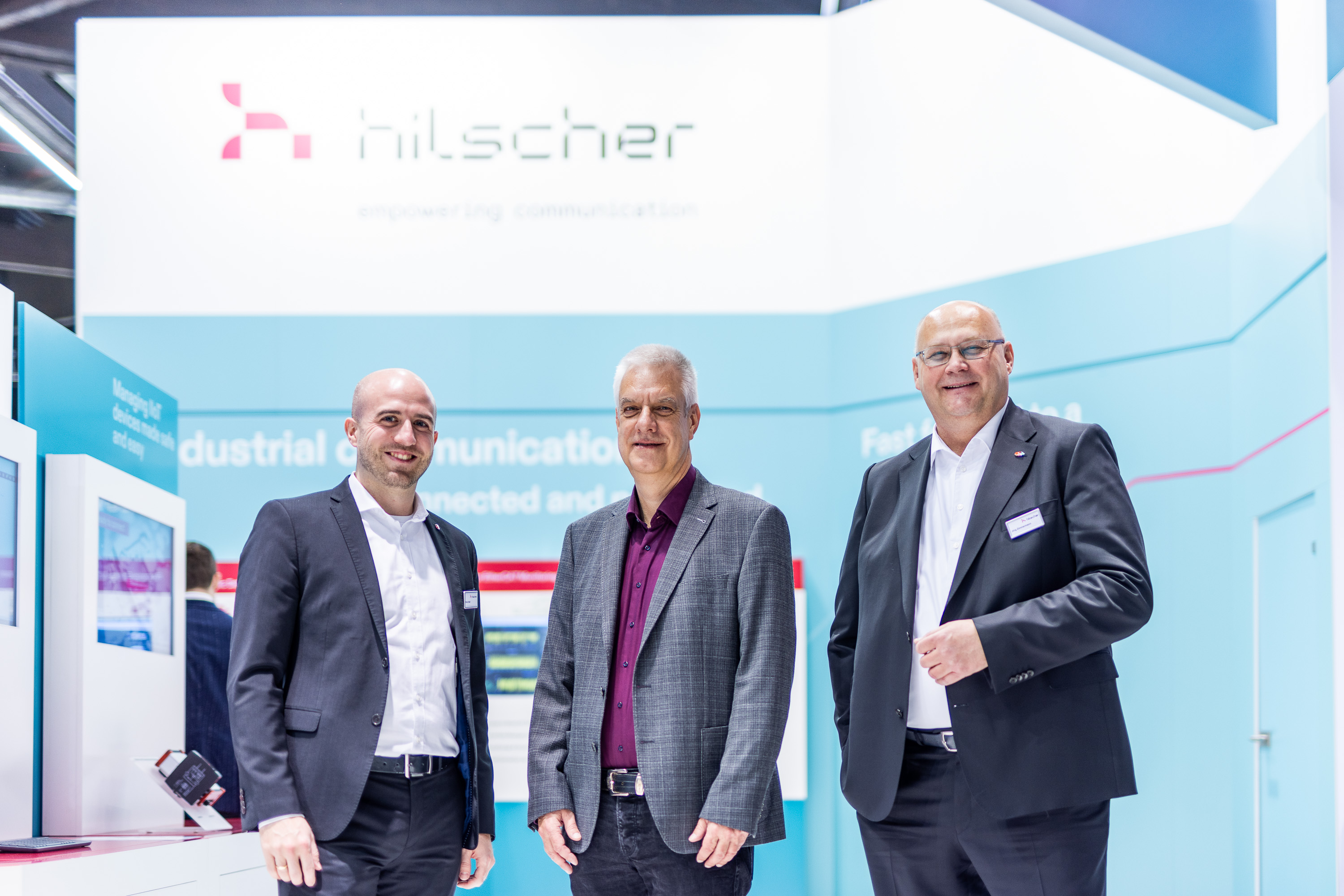 Three men are standing at Hilscher’s trade fair booth at SPS 2023. They are wearing business suits and smile into the camera. In the background a light blue and white wall of the Hilscher booth can be seen with a large Hilscher logo on top.