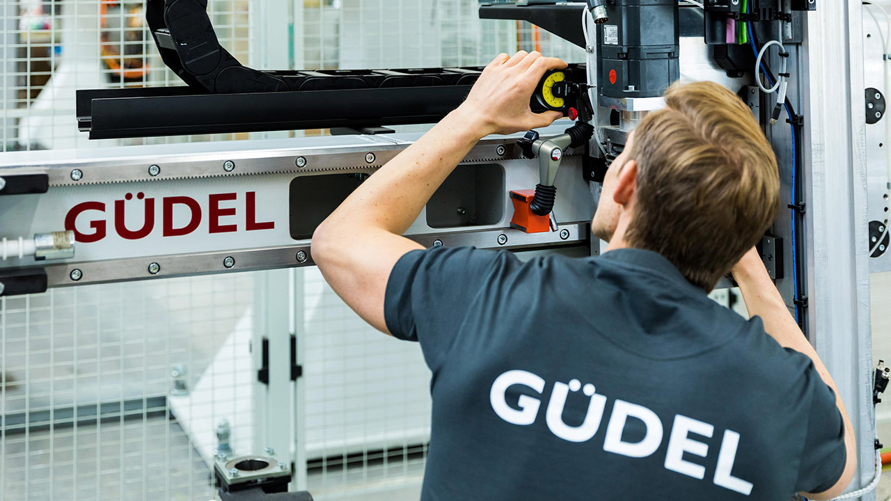 A man inspects the rollers of a drive axis by Güdel. He wears a black shirt with Güdel written in large white letters on it.